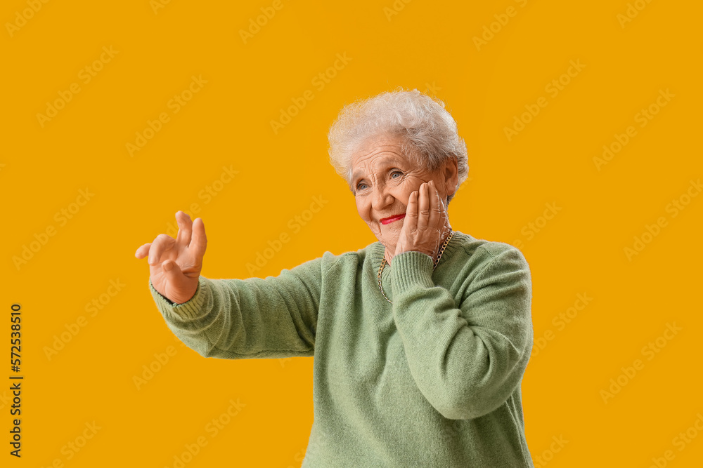 Surprised senior woman in green sweater pointing at something on yellow background