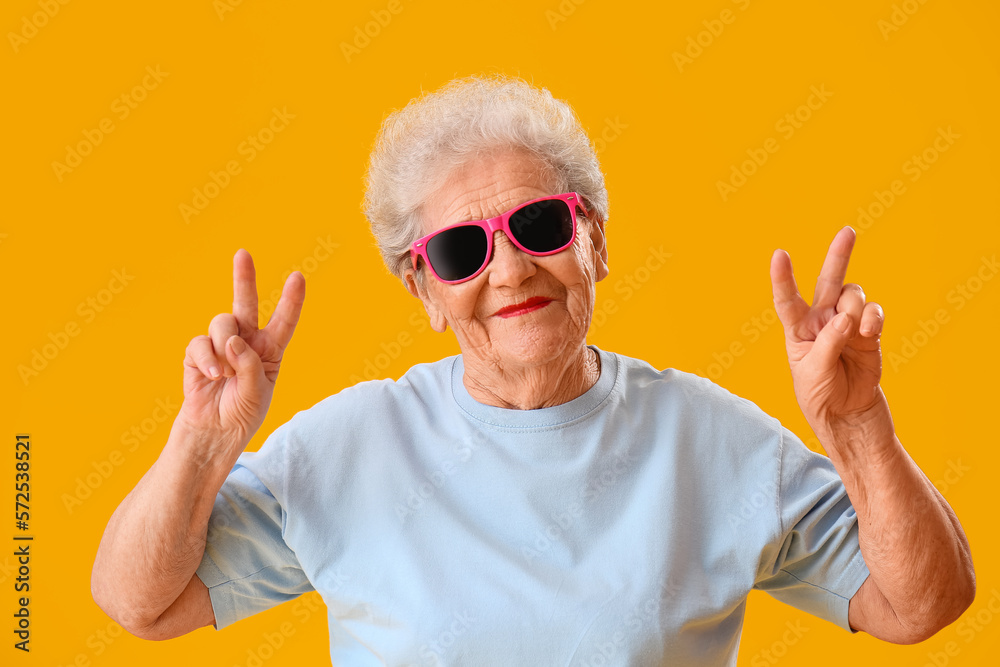 Senior woman in sunglasses showing victory gesture on yellow background