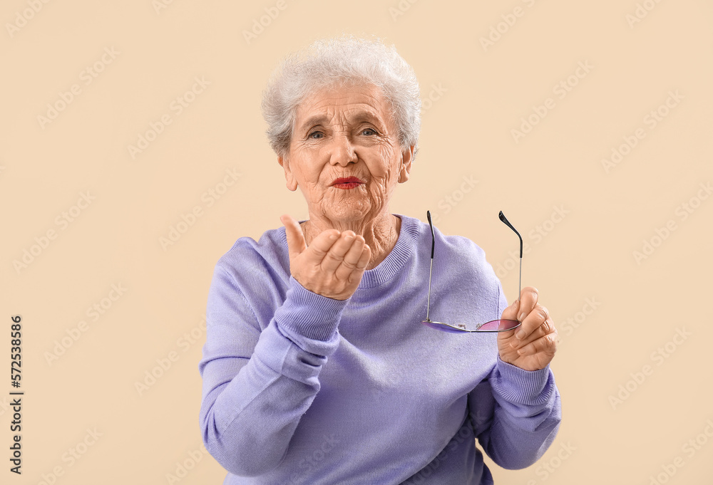 Senior woman in lilac sweater with sunglasses blowing kiss on beige background