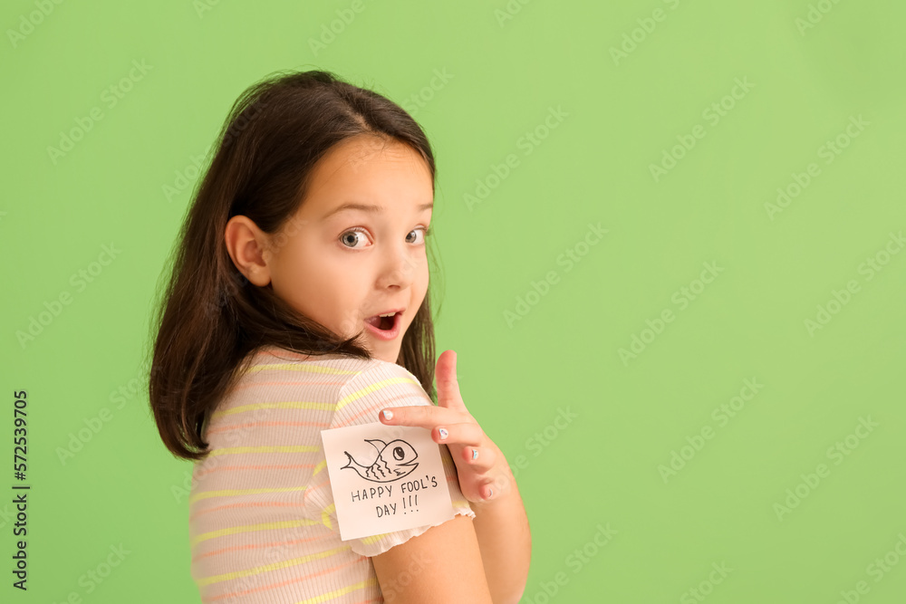 Surprised little girl pointing at sticky paper with text HAPPY FOOLS DAY on her t-shirt against gre