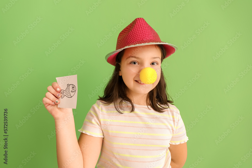 Little girl in funny disguise holding paper with drawn fish on green background. April Fools Day ce