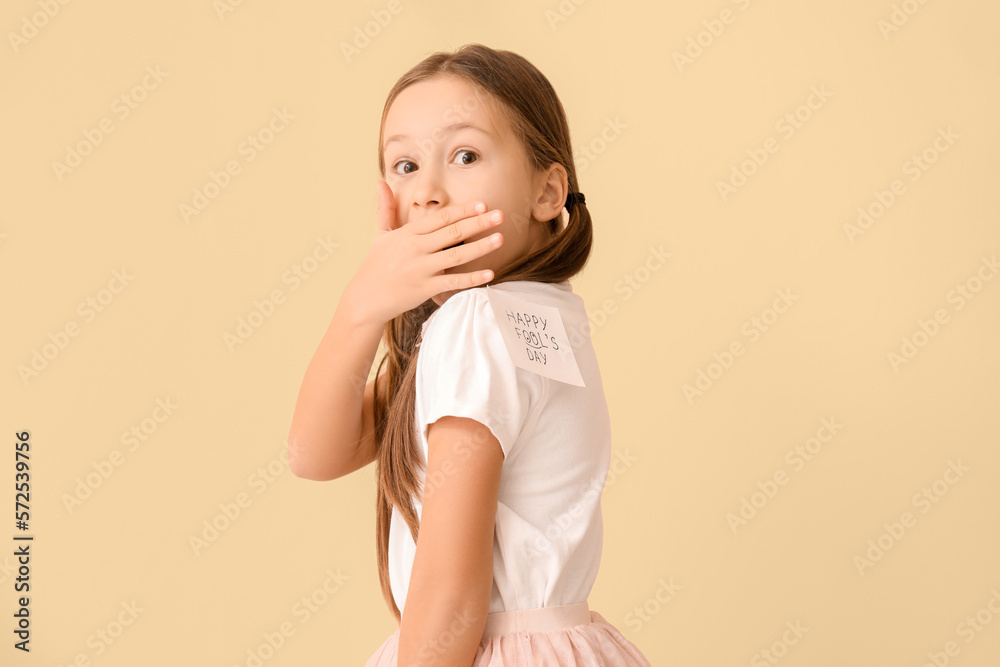 Shocked little girl and sticky paper with text HAPPY FOOLS DAY on her t-shirt against beige backgro