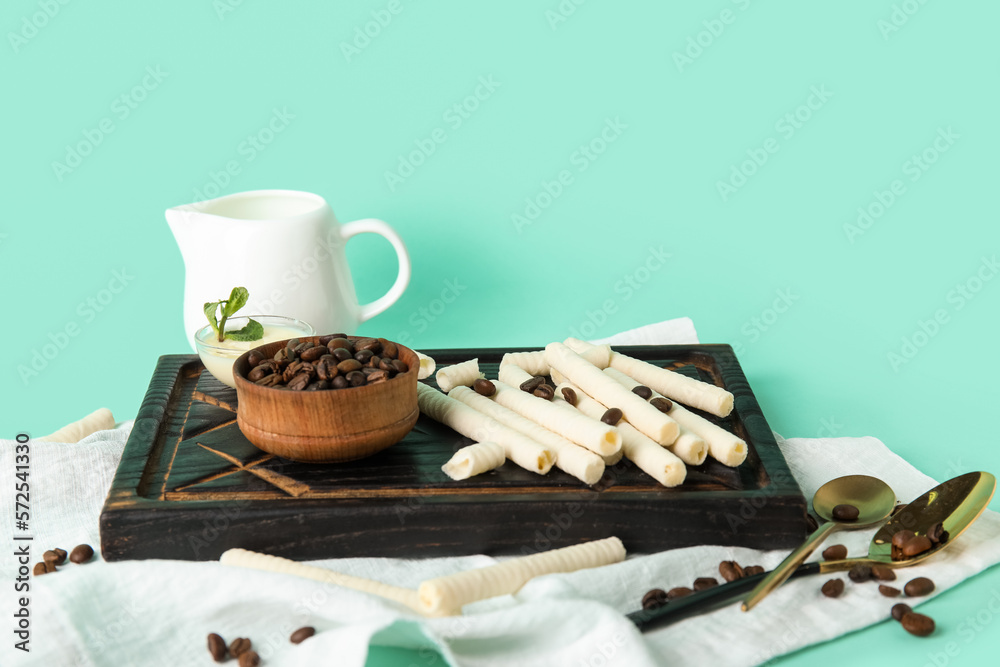 Black wooden board with delicious wafer rolls and bowl of coffee beans on turquoise background