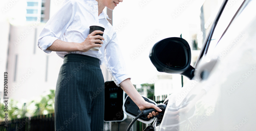 Closeup businesswoman holding coffee, insert EV charger to electric vehicle at public charging stati