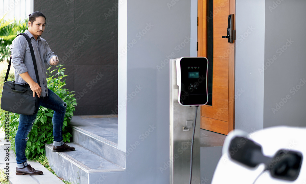 Progressive asian man and electric car with home charging station. Concept of the use of electric ve