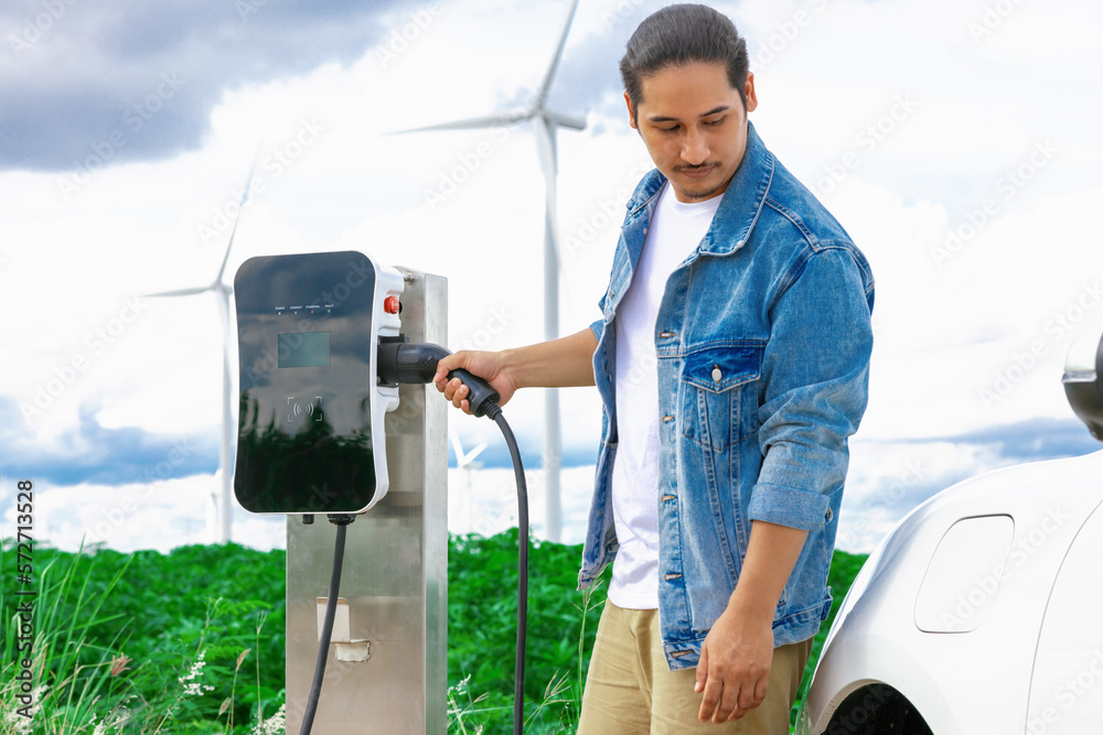 Progressive man with his electric car, EV car recharging energy from charging station on green field