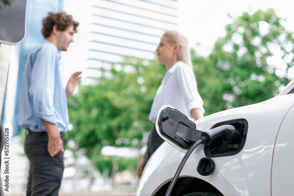 Focus parking-electric car connected to public charging station with blur progressive businesspeople