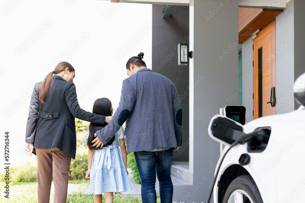 Progressive young parents and daughter with electric vehicle and home charging station. Green and cl