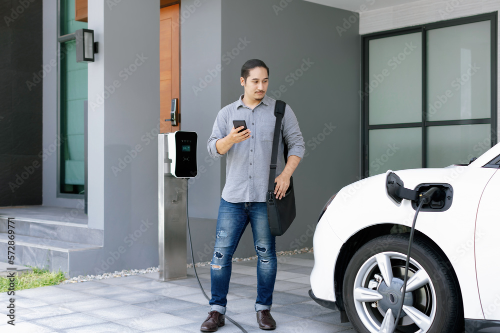 Progressive asian man and electric car with home charging station. Concept of the use of electric ve