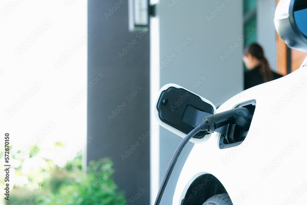 Focus image of electric vehicle recharging battery at home charging station with blurred woman walki