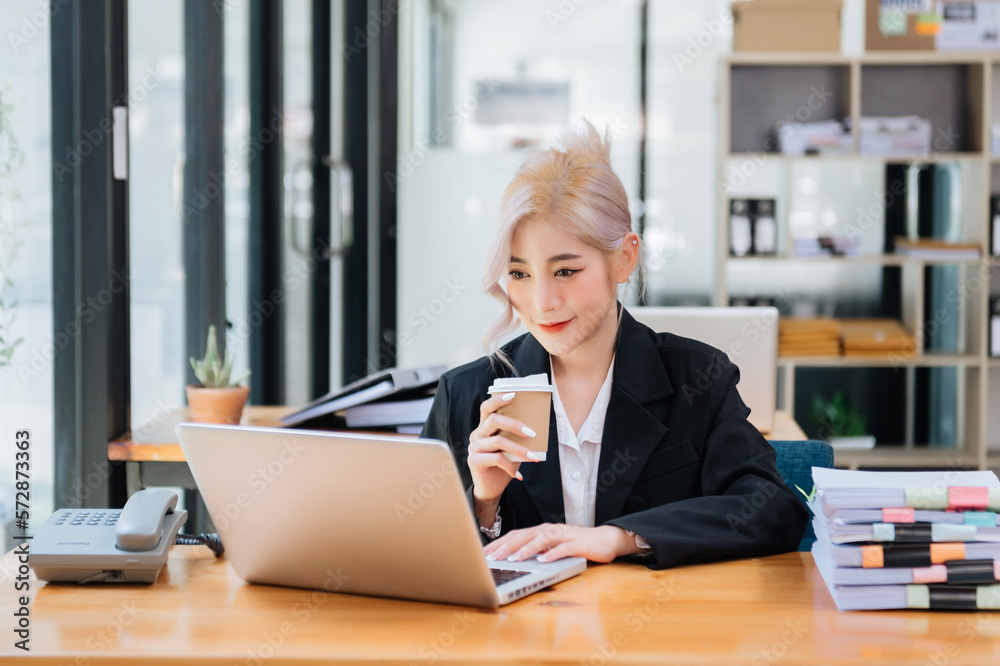 Confident beautiful Asian businesswoman typing laptop computer and digital tablet while holding coff