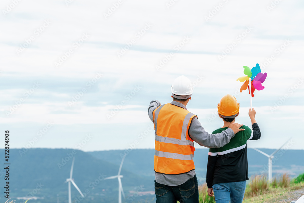 Engineer with his son holding windmill toy on a wind farm atop a hill or mountain. Progressive ideal