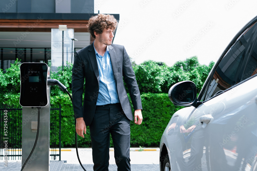 Progressive businessman wearing black suit with electric car recharging at public parking car chargi