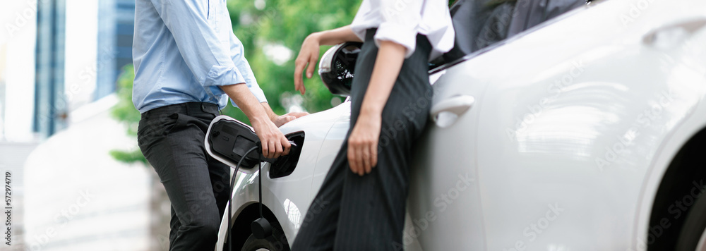 Progressive businessman and businesswoman with electric car connected to charging station before dri