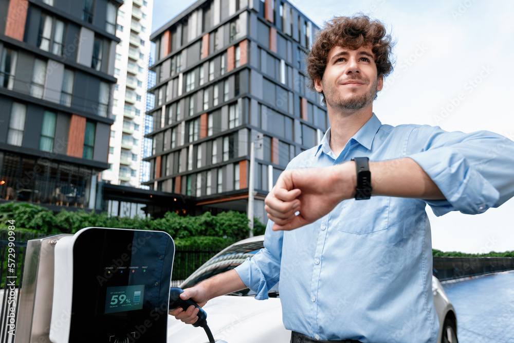 Businessman with smartwatch at modern charging station for electric vehicle with background of resid