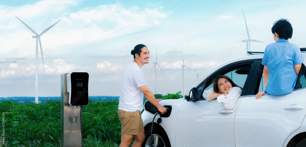 Concept of progressive happy family enjoying their time at wind farm with electric vehicle. Electric