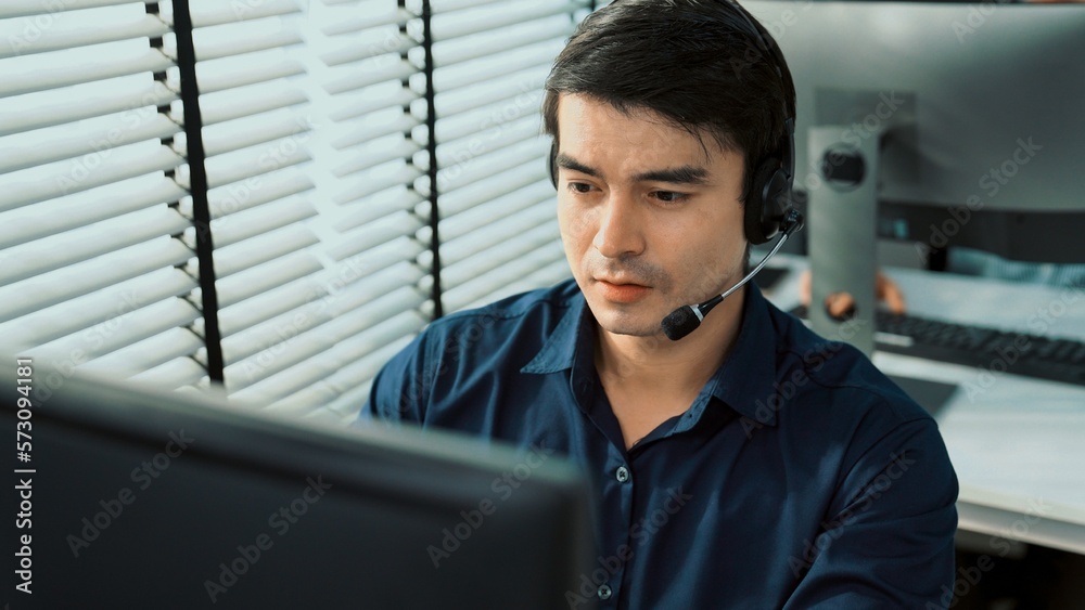 Young competent asian male call center agent working at his computer while simultaneously speaking w