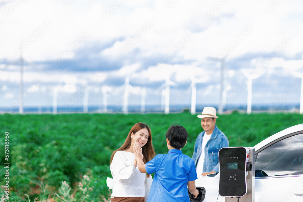 Concept of progressive happy family enjoying their time at wind farm with electric vehicle. Electric