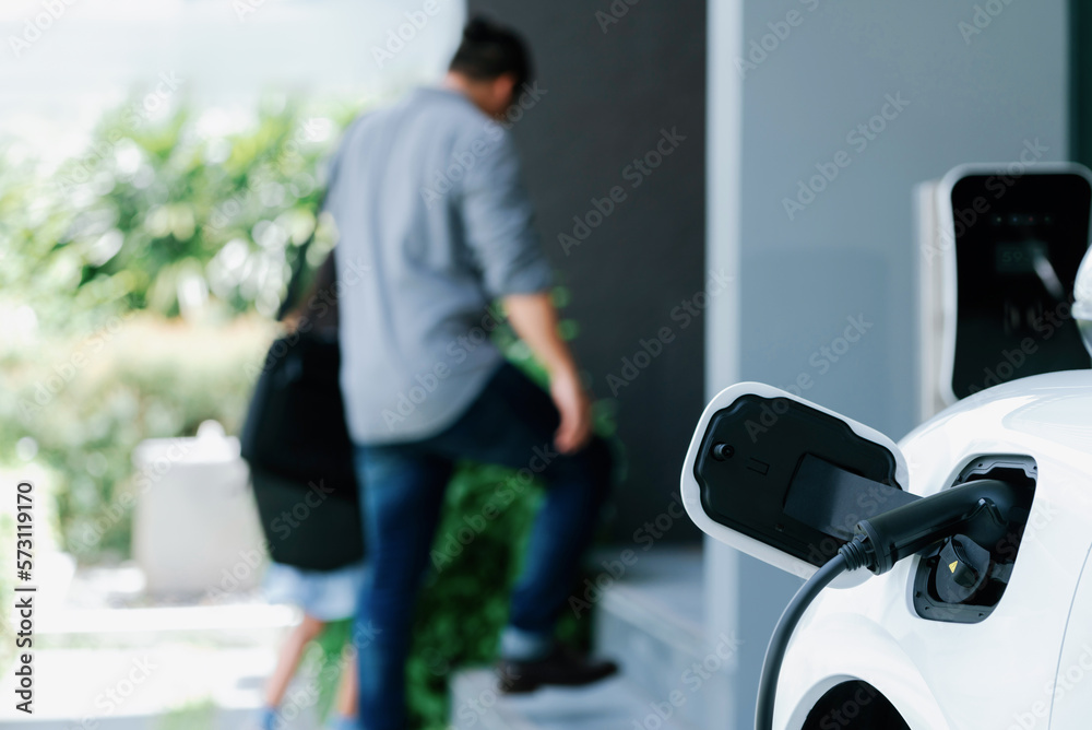 Focus electric car recharging at home charging station with blurred father and daughter walking in b