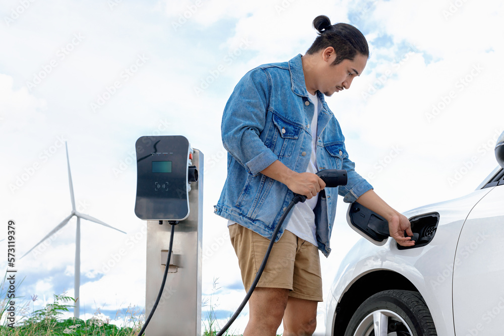 Progressive man with his electric car, EV car recharging energy from charging station on green field