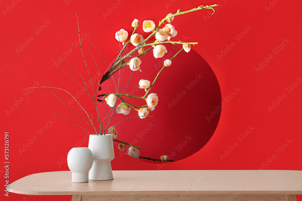 Vases on wooden table near red wall with blooming Chinese plum branches in room