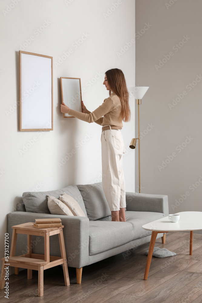 Young woman hanging blank frame on light wall at home