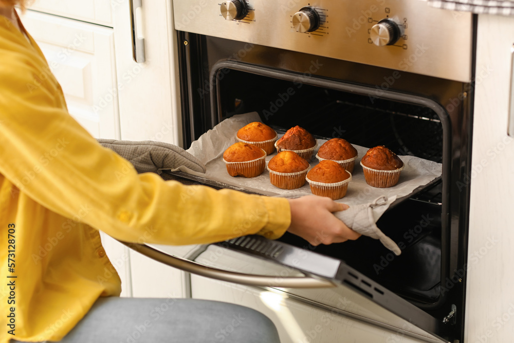 Young woman taking tray with cupcakes from oven in kitchen, closeup