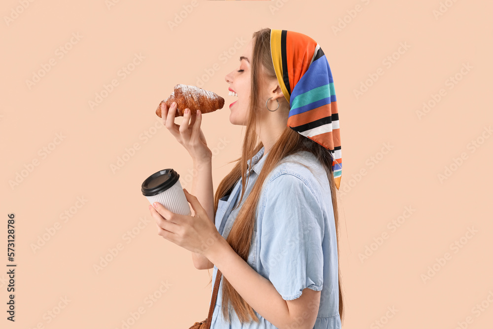 Young woman with cup of coffee eating croissant on beige background