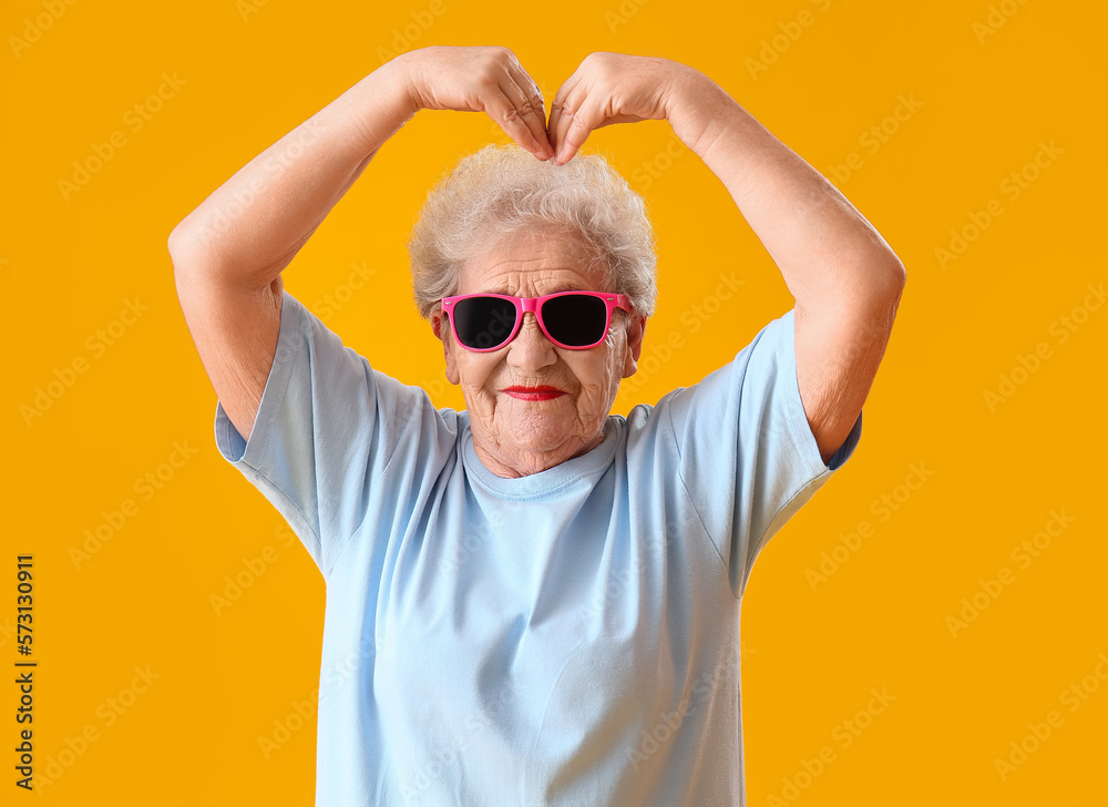 Senior woman in sunglasses making heart shape with hands on yellow background