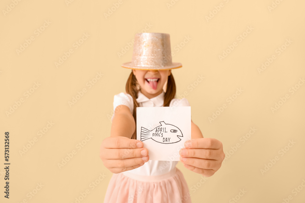 Little girl in hat holding paper with drawn fish on beige background, closeup. April Fools Day cele