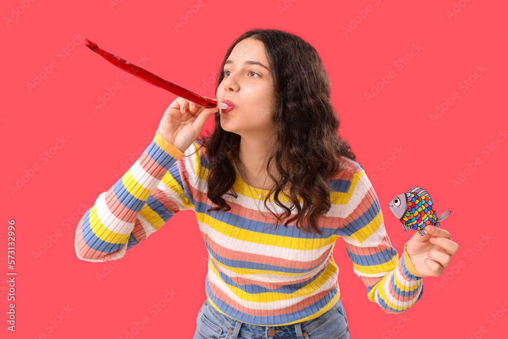 Teenage girl with party blower and paper fish on red background. April Fools Day celebration