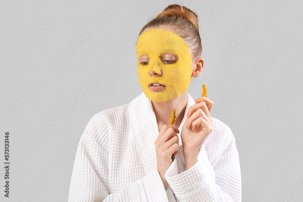 Young woman in bathrobe with turmeric sheet mask and roots on grey background