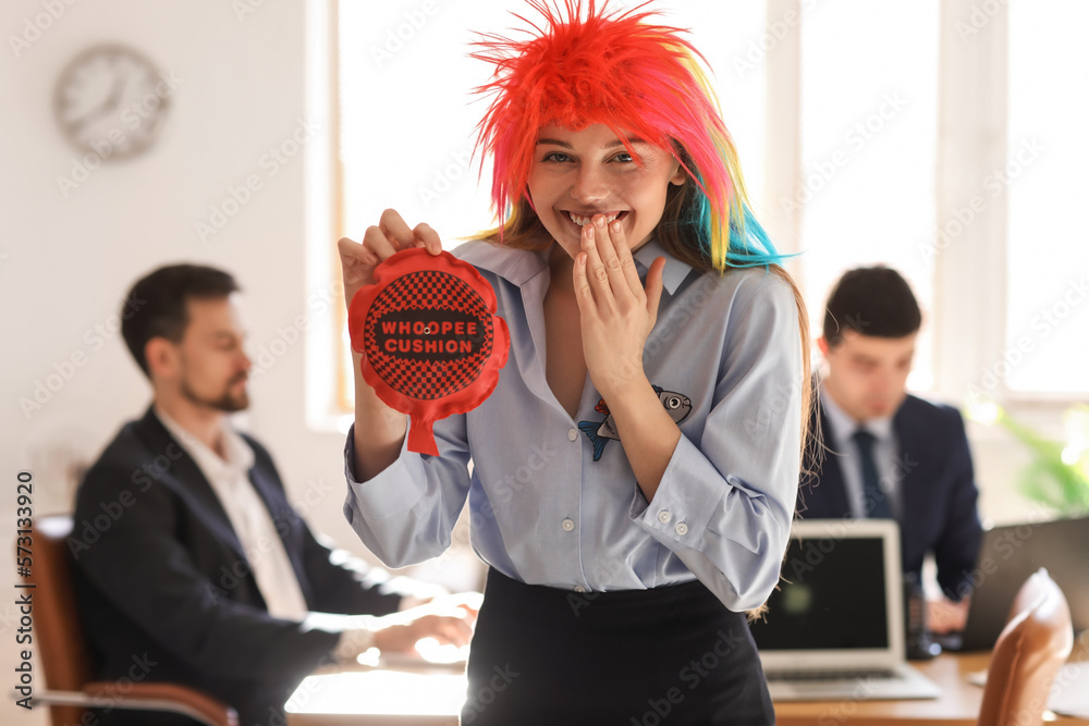 Young woman with funny wig and whoopee cushion in office. April Fools Day celebration