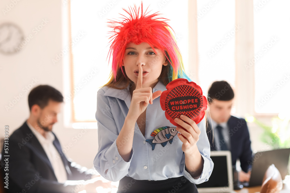 Young woman with funny wig and whoopee cushion showing silence gesture in office. April Fools Day c