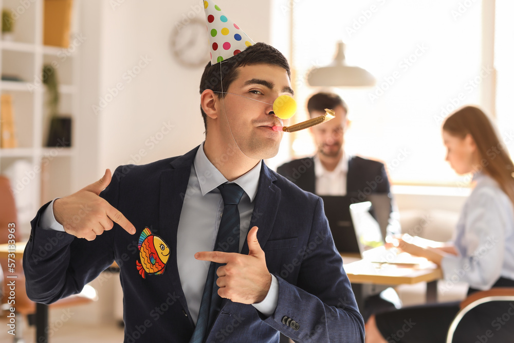 Young man with party blower and clown nose pointing at paper fish in office. April Fools Day celebr