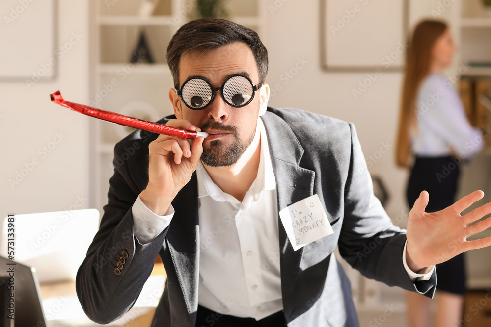 Young man with funny eyeglasses and party blower in office. April Fools Day celebration