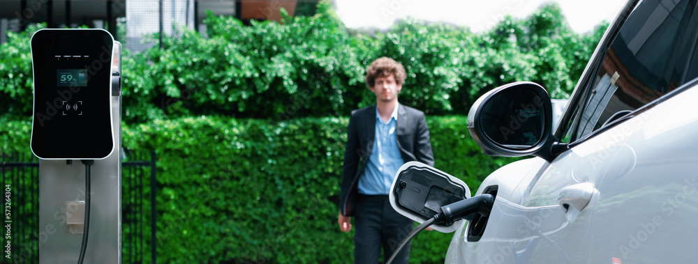 Progressive businessman wearing black suit with electric car recharging at public parking car chargi