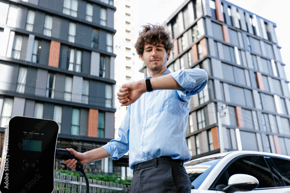 Businessman with smartwatch at modern charging station for electric vehicle with background of resid