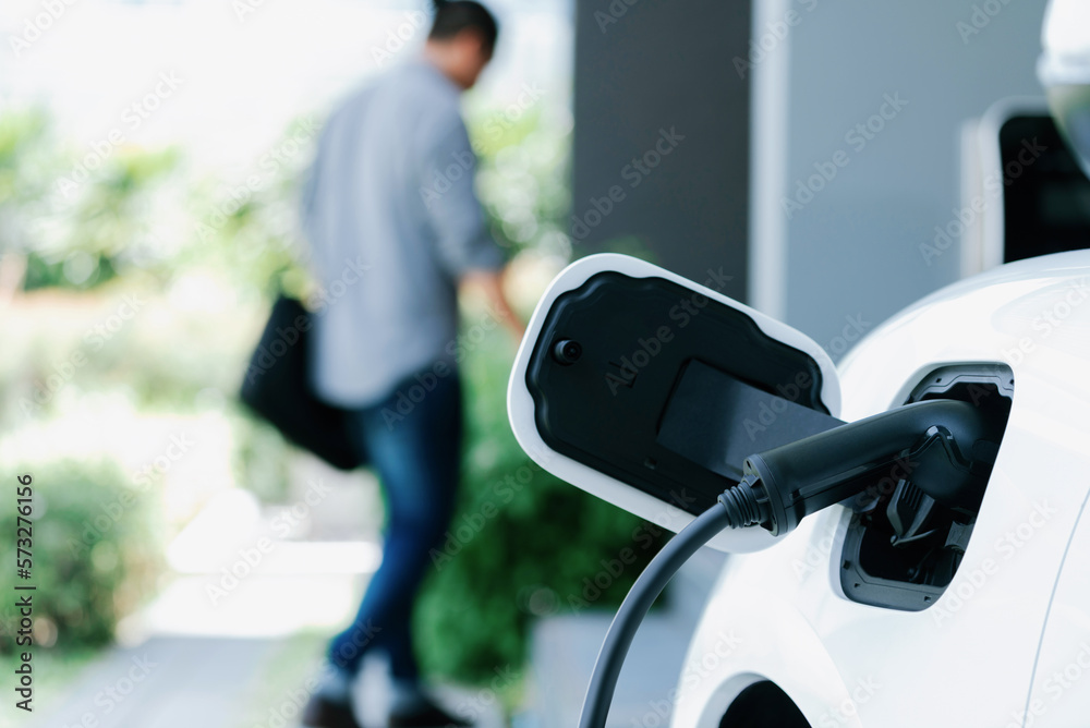 Focus electric car charging at home charging station with blurred progressive man walking in the bac