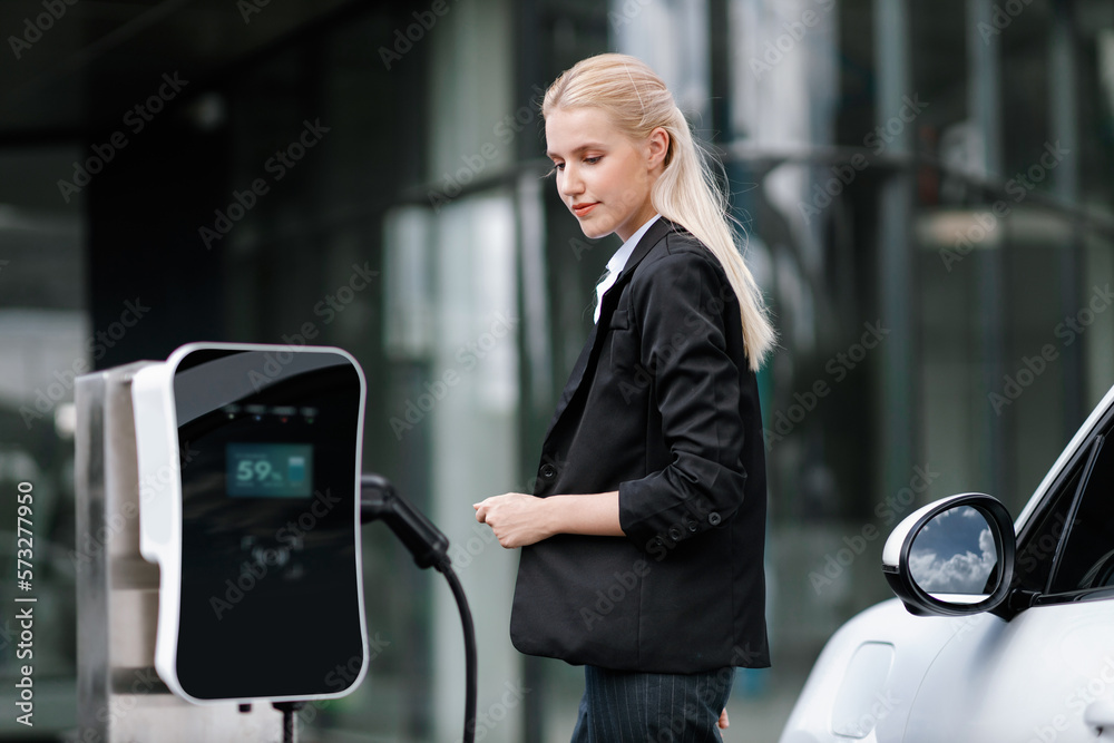 Progressive businesswoman insert charger plug from charging station to her electric vehicle with apa