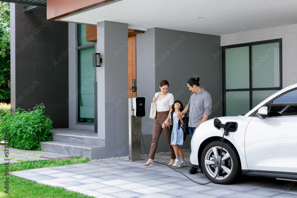 Progressive young parents and daughter with electric vehicle and home charging station. Green and cl