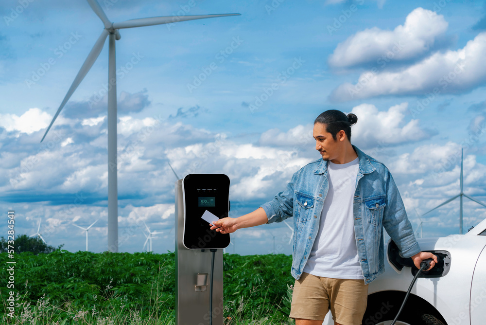 Progressive man with his electric car, EV car recharging energy from charging station on green field