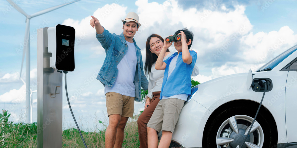 Concept of progressive happy family enjoying their time at wind farm with electric vehicle. Electric