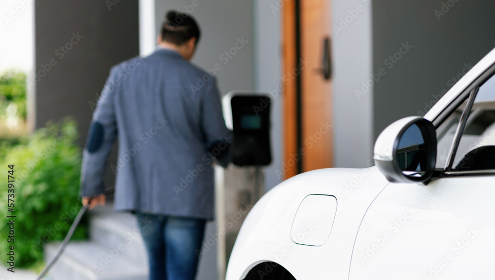 Focus electric car charging at home charging station with blurred progressive man walking in the bac