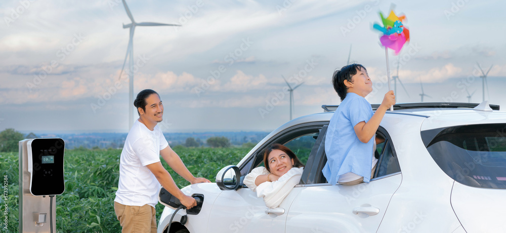 Concept of progressive happy family enjoying their time at wind farm with electric vehicle. Electric