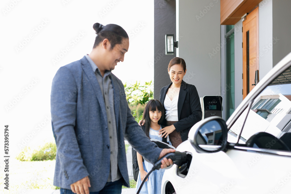 Progressive young parents and daughter with electric vehicle and home charging station. Green and cl