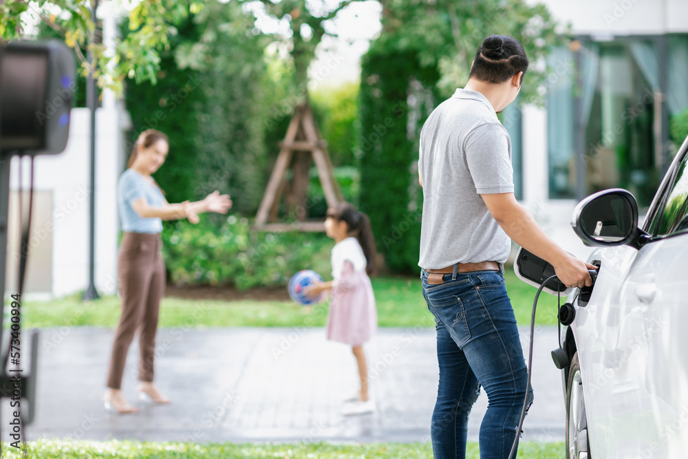 Focus image of progressive man charging electric car from home charging station with blur mother and