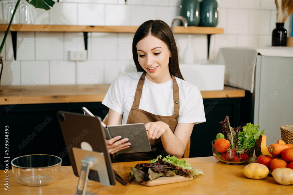 Young beautiful woman in the kitchen in an apron, fresh vegetables on the table, writes down her fav