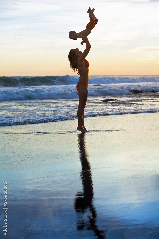 Happy family swimming fun on sea beach - mother tossing up baby son into mid air, catching on sunset