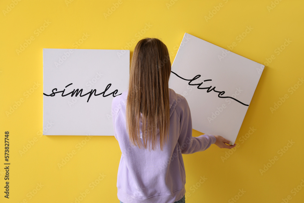 Young woman hanging poster on yellow wall at home